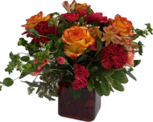 Floral arrangement with orange roses, red and bright pink carnations in a ruby colored square vase.